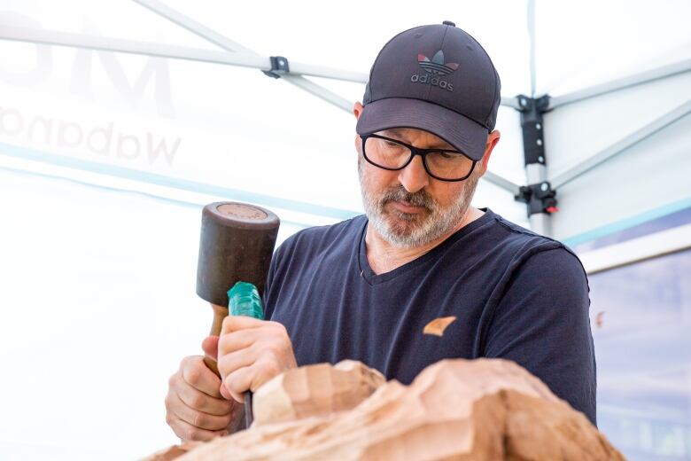 Man in glasses and baseball cap holds a carving tool and chisels a piece of wood.