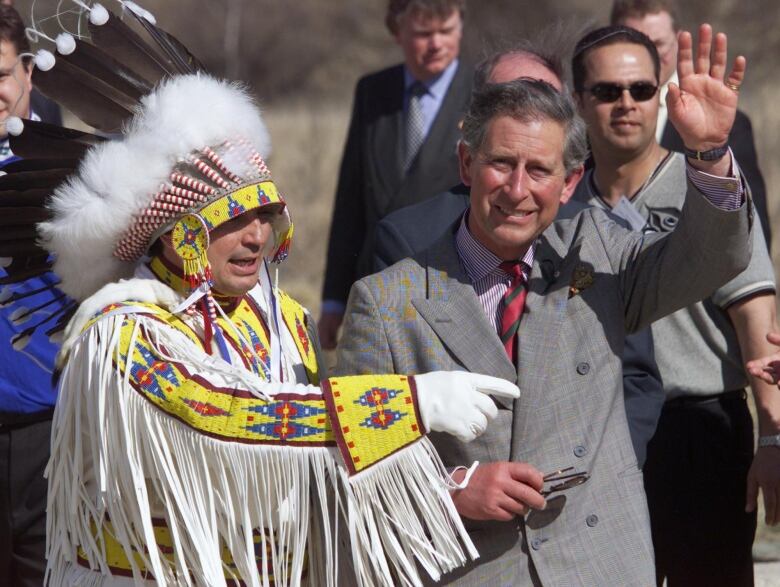 A First Nations Chief in ceremonial garb speaks and points off-camera to the right as he stands next to a smiling man in a grey suit lifting one hand up to wave.