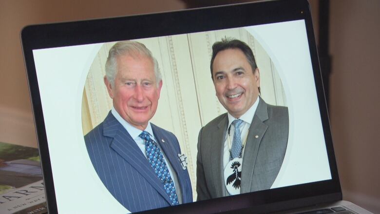 A photo of two smiling men is shown on a tablet screen: at left, one man wears a blue pinstriped suit and blue tie, the other wears a grey suit, blue tie and a medallion.