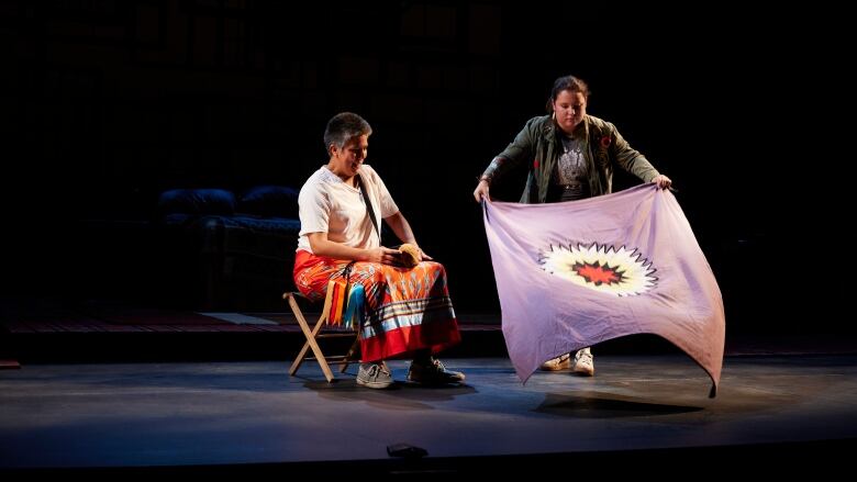 Salt Baby (Chanin Lee) is laying out a First Nations blanket in front of an elder (Nicole Joy-Fraser) who sits nearby.