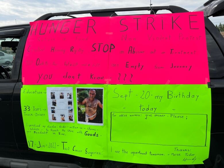 A pink and neon yellow sign are taped on to a car. In part, it reads, 'Hunger Strike. Refuse of food. Non-violent protest. Central Housing Registry stop an abusive act or treatment.'