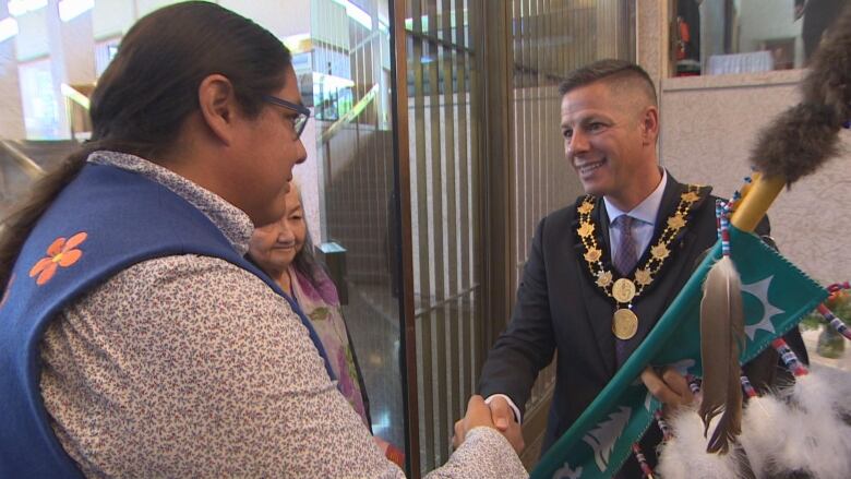 A man wearing a ceremonial chain and holding an Indigenous ceremonial staff, decorated with feathers, shakes the hand of another man, wearing a blue vest and long dark hair, tied back in a pony tail.