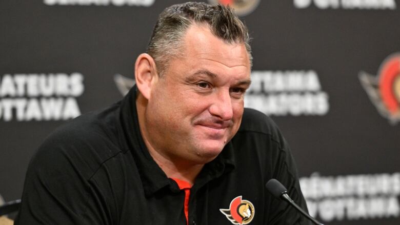 A man in an Ottawa Senators golf shirt in front of a microphone with more team logos in the back.