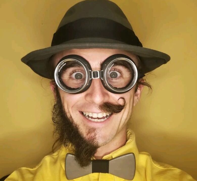 A man with a beard on the right side of his face and a curly moustache on the left wears a fedora hat, large round glasses, a solid bowtie and a bright yellow shirt. He stands in front of a yellow background and smiles excitedly.