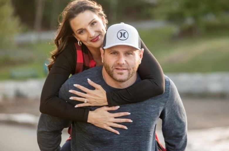 A man with a Hurley brand baseball cap on is seen giving a piggy-back ride to a woman with long hair. They're both looking at the camera.