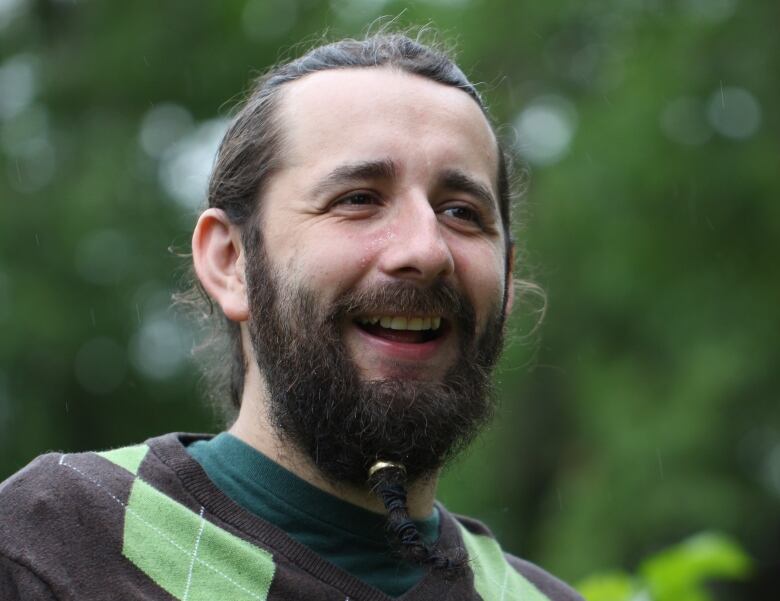 A man with long, brown hair and a beard in a ring smiles. He is wearing an argyle sweater and standing in front of a lush, green background that is blurred.