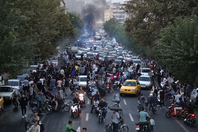 A crowd, including people, scooters, motorcycles and vehicles, blocks a road.