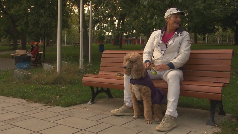 a man wearing a beige baseball cap and jacket with a red shirt and white pants sits on a bench with his dog 