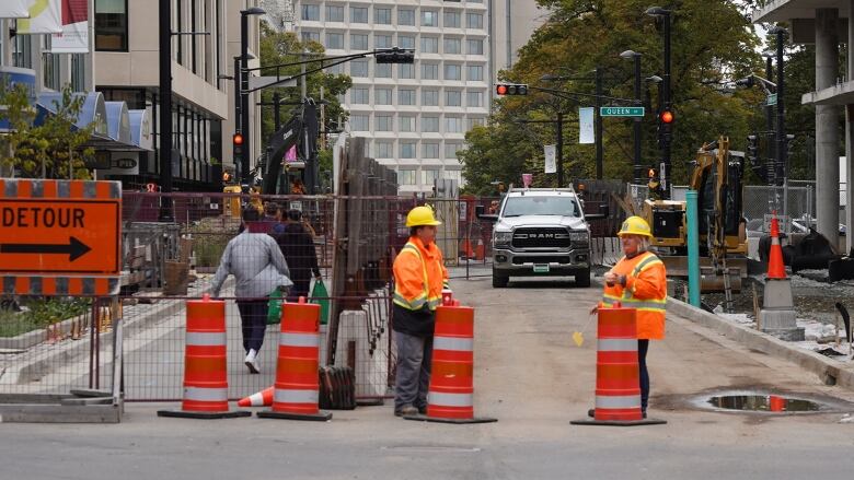 Construction workers in orange and yellow jackets and hats stand behind orange cones blocking part of Spring Garden. Red fencing is up beside them.