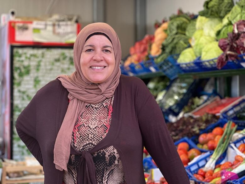 A woman in a head scarf smiles. She is standing in front of a fruit stand.