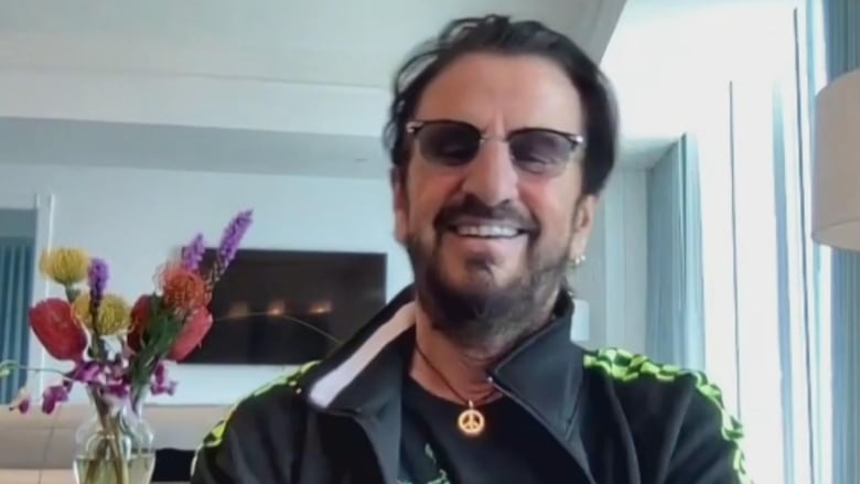 A bearded man in sunglasses, wearing a peace symbol necklace, smiles at the camera.