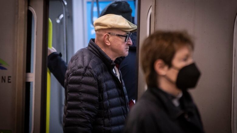 An unmasked man enters a SkyTrain, with a masked person visible in the foreground.