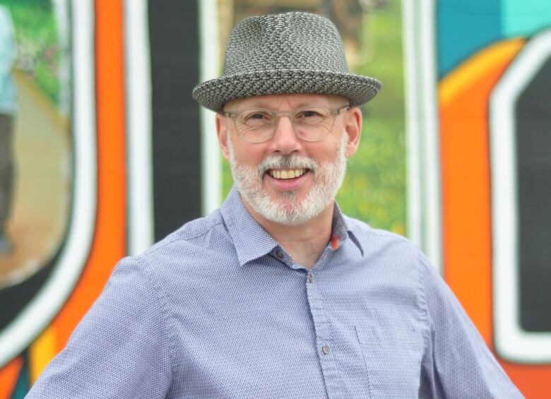 A man in a fedora with a white beard smiles and stands with his hands on his hips in front of a colourful background.