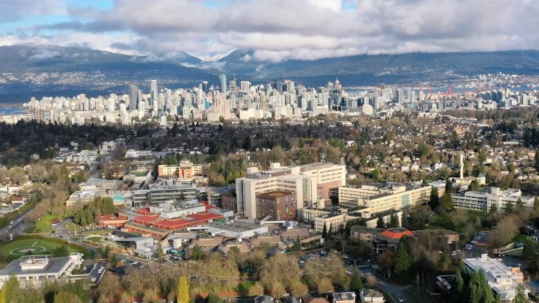 An aerial photo shows a hospital campus surrounded by trees and single-family homes, with the Vancouver skyline, English Bay and the North Shore mountains in the background.