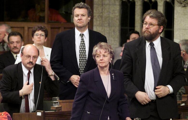 A woman in a blue blazer stands in the House of Commons, with three men standing behind her.