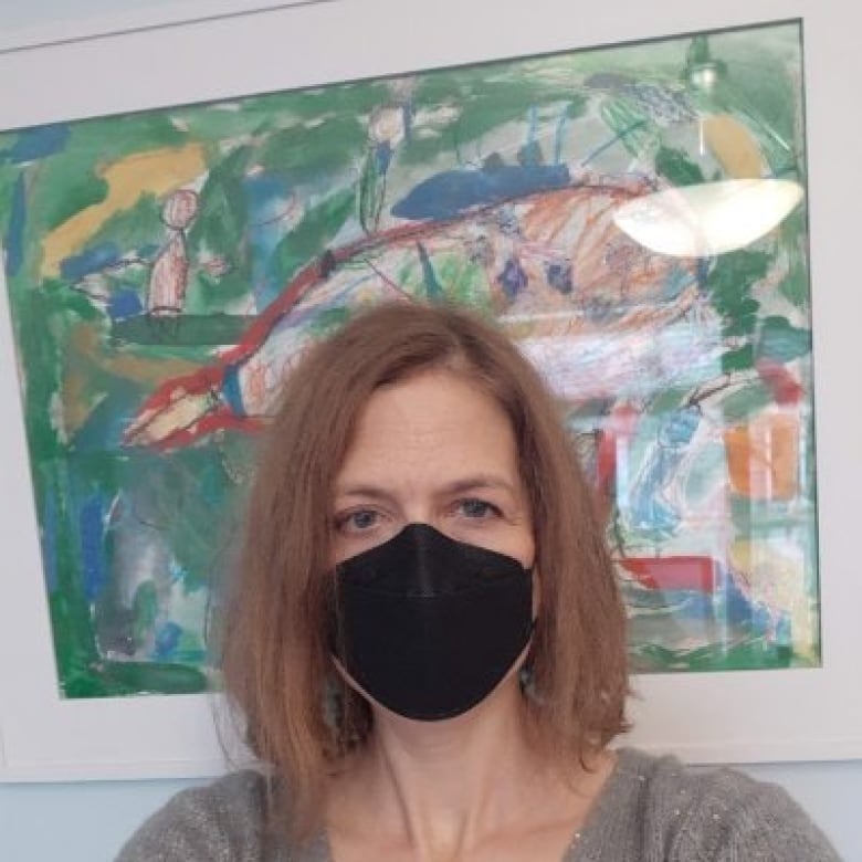 A brown-haired woman wears a black mask in front of a drawing.