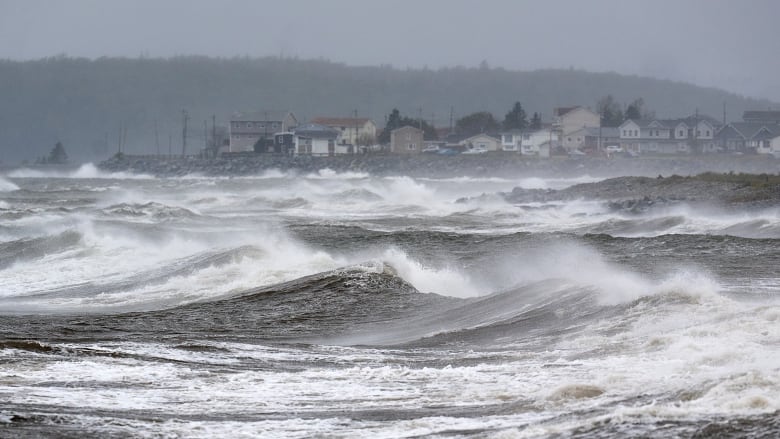 Waves and sea spray can be seen with a line of houses in the distance.