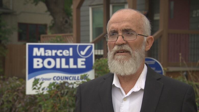 A man with glasses and a white beard wears a suit and stands in front of a council election sign.