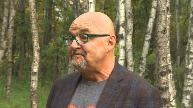 A bald man in green glasses smiles in front of a forest.