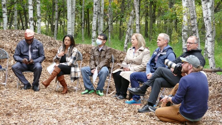 A group of six people sit in chairs on wood chips with a forest behind them. A person with a camera crouches next to them.