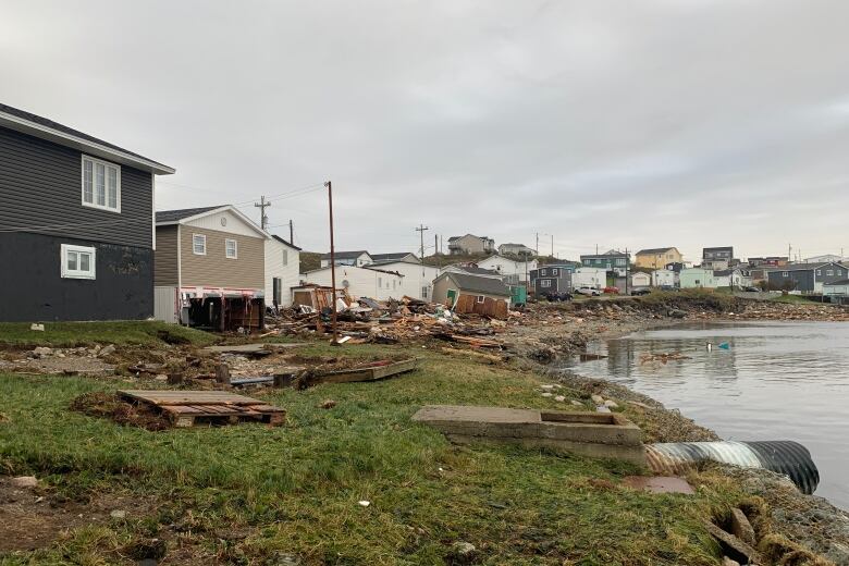 Houses line a grassy shore. The shore is scattered with debris and some of the homes have been damaged, and buildings have been knocked off their foundations.