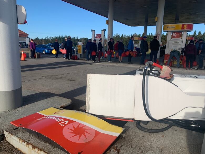 A gas pump lies on the ground as people line up at another pump to fill jerry cans for their generators.