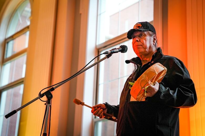 A person in baseball hat and black jacket stands at a microphone about to sing, holding a drum.