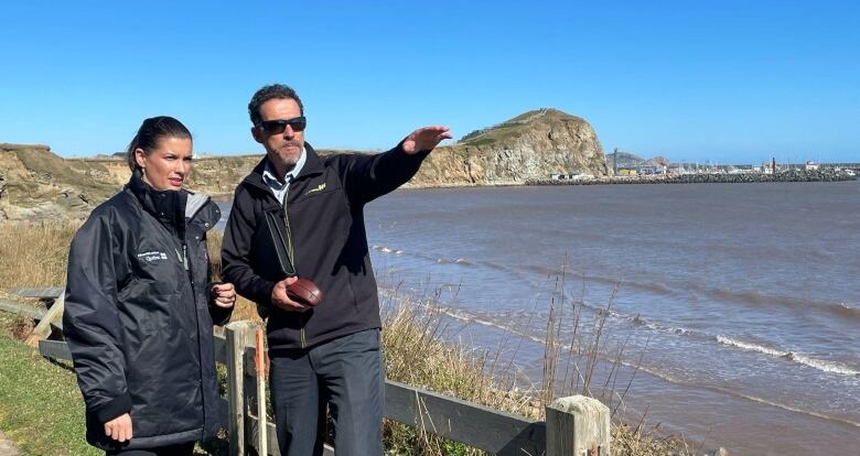 A woman and a man standing near a beach. The man is pointing at something that is not visible in the picture.