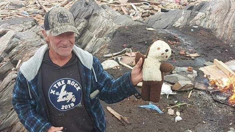 A man stands on a beach holding a weathered teddy bear, with wooden debris strewn around him.