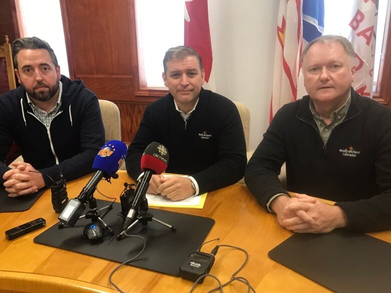 Three men sit a desk for a press conference. Two microphones sit in the middle of the table, and flags of Newfoundland and Labrador and Canada stand behind them.