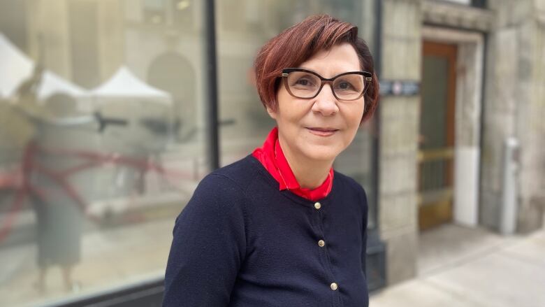 A middle-aged woman with glasses stands on a city sidewalk and smiles.