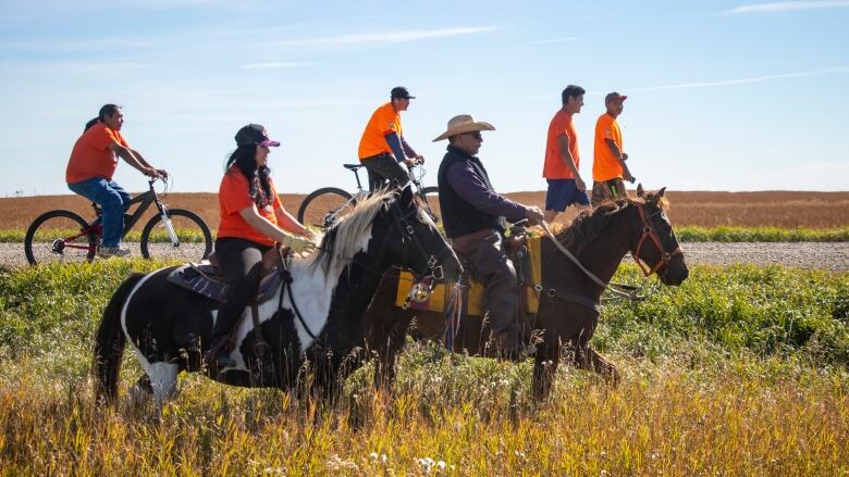 Two people ride horses while people behind them walk and ride bikes. Everyone is wearing orange shirts.