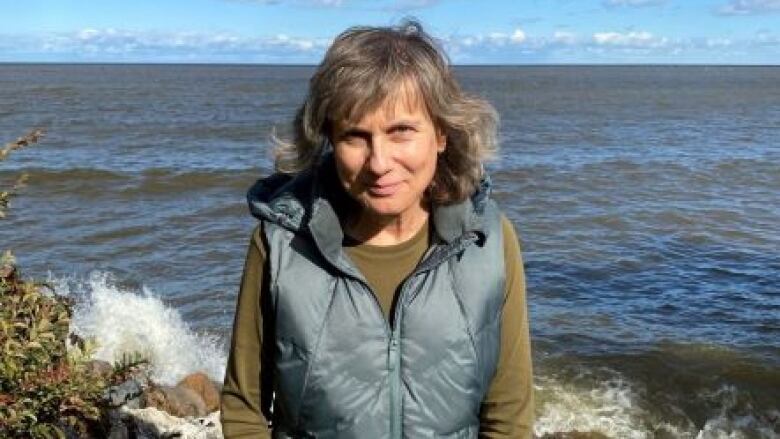 A woman with streaked gray hair stares straight ahead, standing on a rocky shoreline in front of a lake.