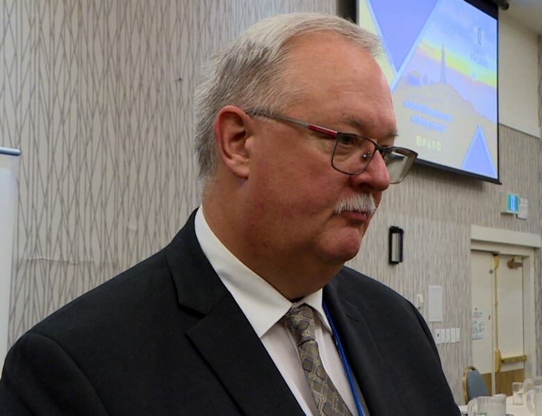 A head and shoulders shot of a person, seen from the side, wearing with white hair and a mustache wearing glasses.