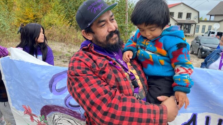 A bearded man in a red plaid shirt holds a little boy in his arms.