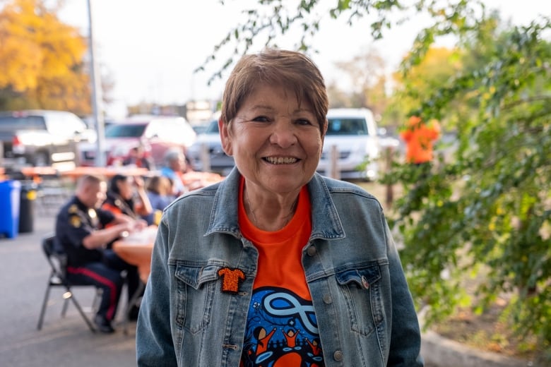 A woman stands for a medium-close portrait. She is wearing a jean jacket and an orange 'every child matters' shirt, with a beaded orange shirt pin attached to her jacket. 