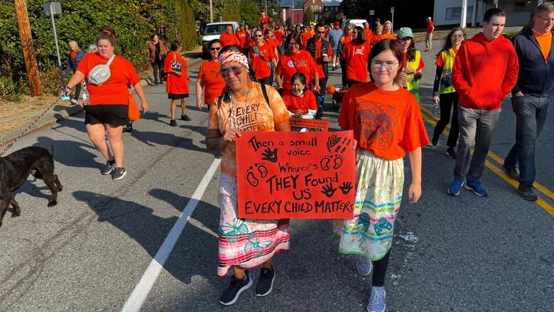 Groups of people marching wearing orange garments. Someone at the front of the line has a sign that reads 'Then a small voice whispers They Found Us Every Child Matters'.