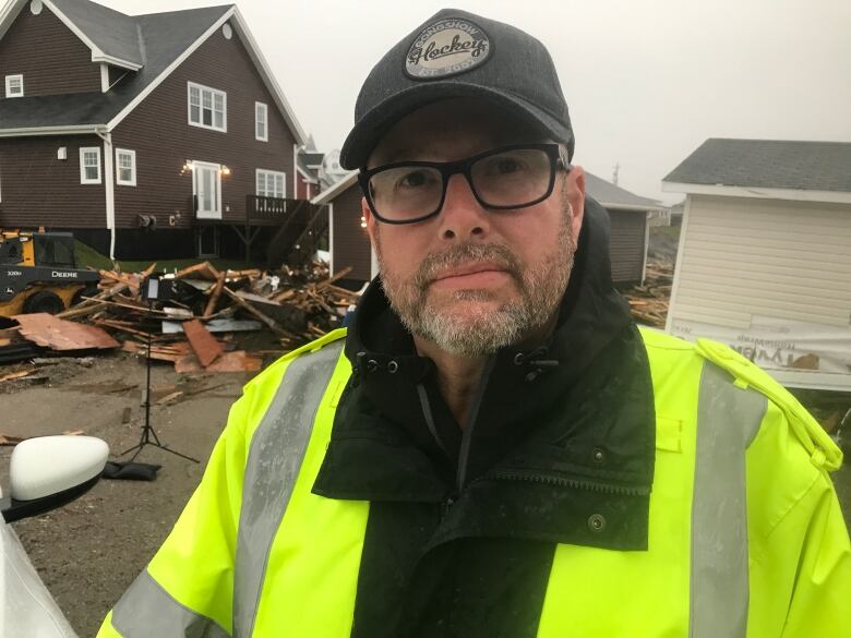 A head and shoulders shot of a person wearing a black hat, glasses and a bright yellow safety jacket. Behind the man, to the left, remnants of a structure can be seen.