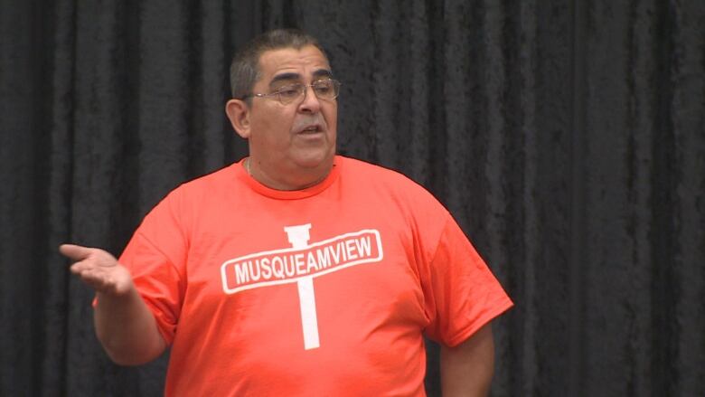 A brown man wears an orange shirt with the word 'Musqueamview' on a street sign.