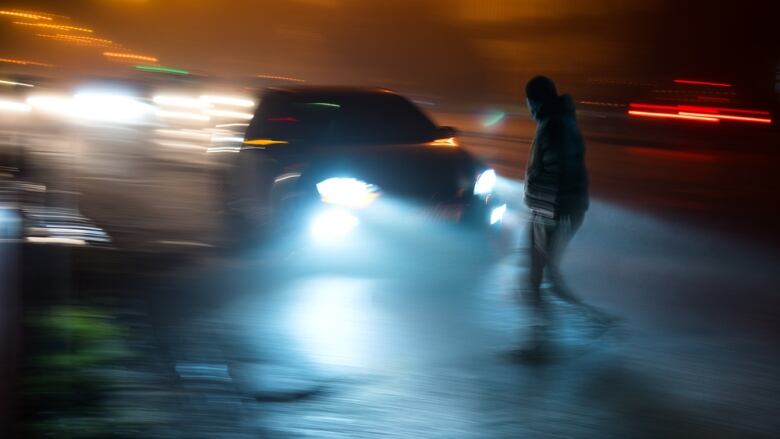 A blurred nighttime photo shows the headlights of a speeding car approaching a pedestrian walking across the street.