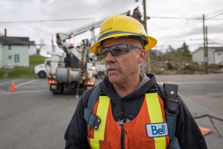 A man wearing a yellow hard hat with Bell Aliant written on it is seen. He is also wearing sunglasses and a neon orange vest over a hoodie.