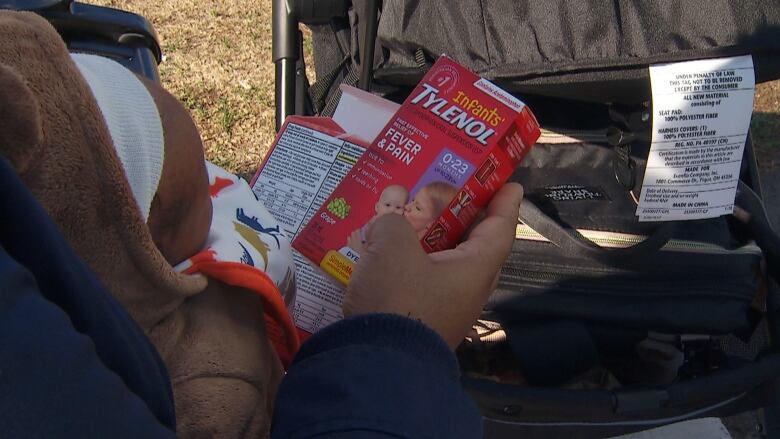 A woman holds a baby and box of Infants' Tylenol by a stroller.