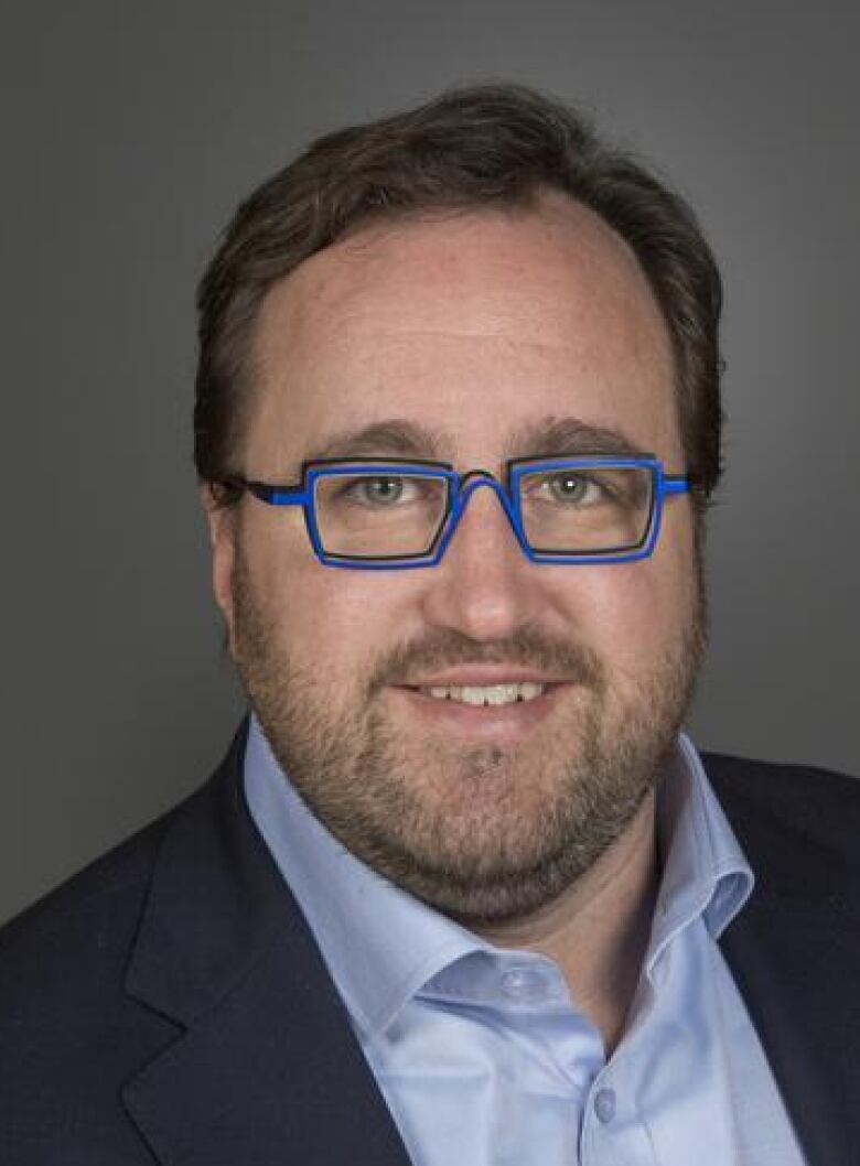 A close-up of a middle-aged man wearing fashionable blue glasses and a dress shirt with an open collar.