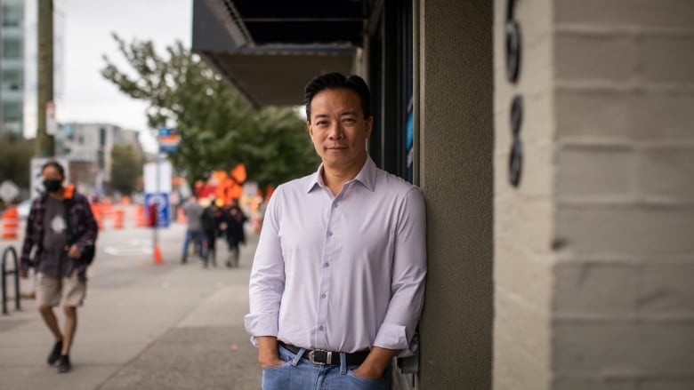 An East Asian man leans against a wall. He is wearing a light purple shirt.