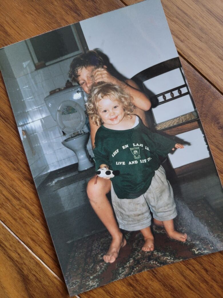 A child with curly blond hair looks at the camera while a slightly older boy plays with her hair behind her. 