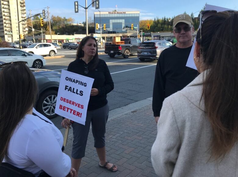 Woman stands on sidewalk holding protest sign, surrounded by several other protestors.