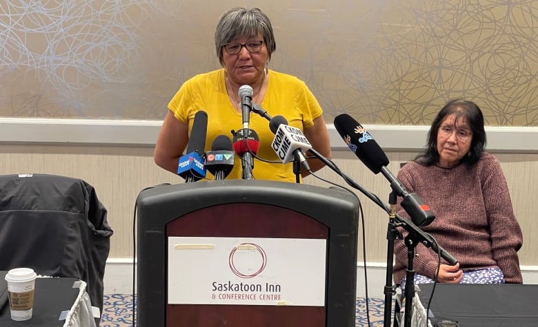 A woman in a yellow T-shirt speaks at a podium in a conference room.