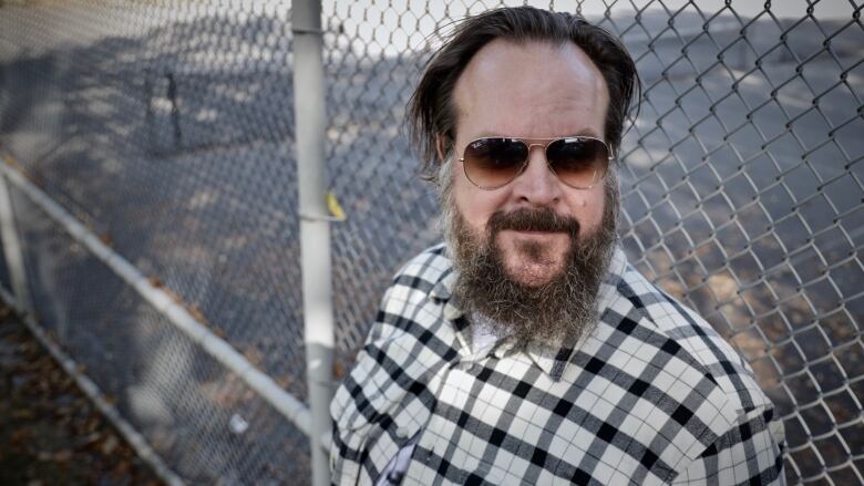 A white man with a beard and sunglasses smiles in front of a chain link fence.