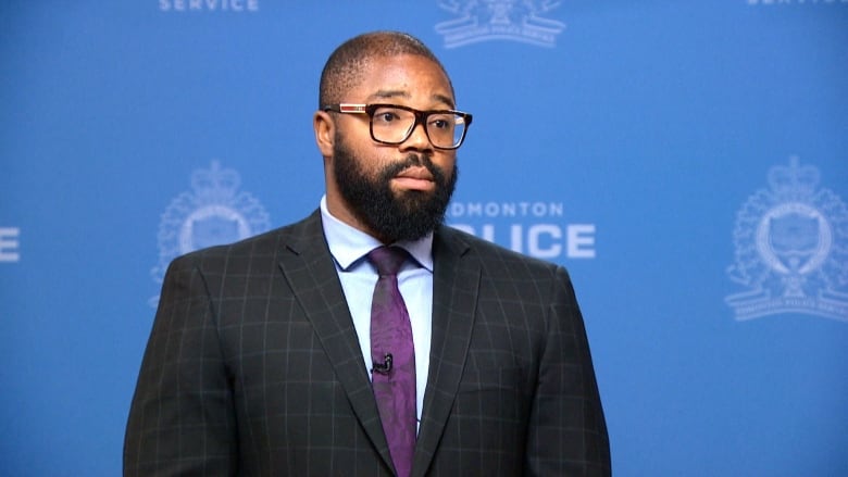 A man in a plaid suit, with shaved head and beard, stands before a blue background