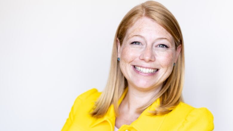 A woman with red hair smiles at the camera. She is wearing a yellow blazer.